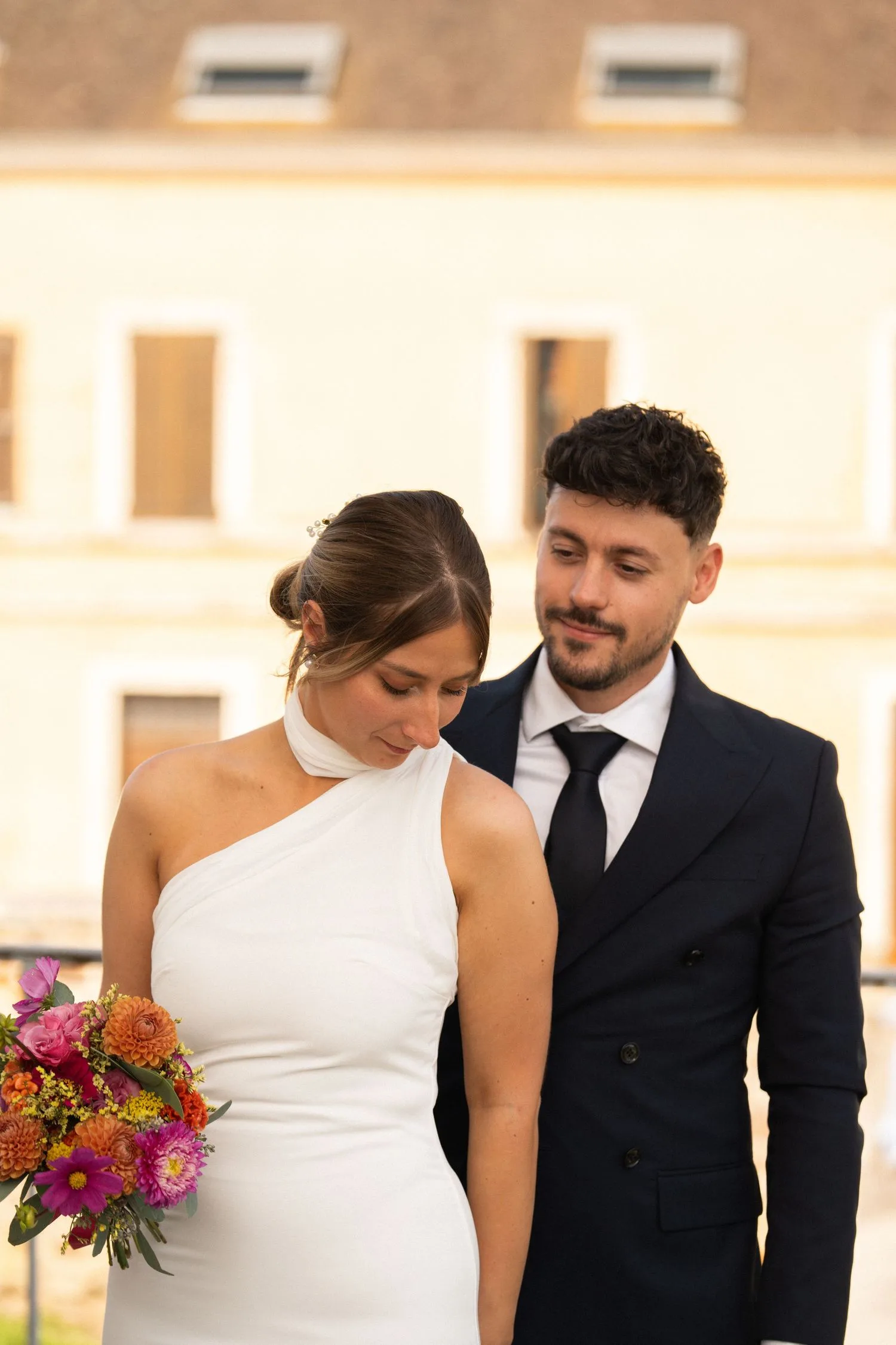 Les mariés devant une grande bâtisse au coucher du soleil, bouquet coloré à la main